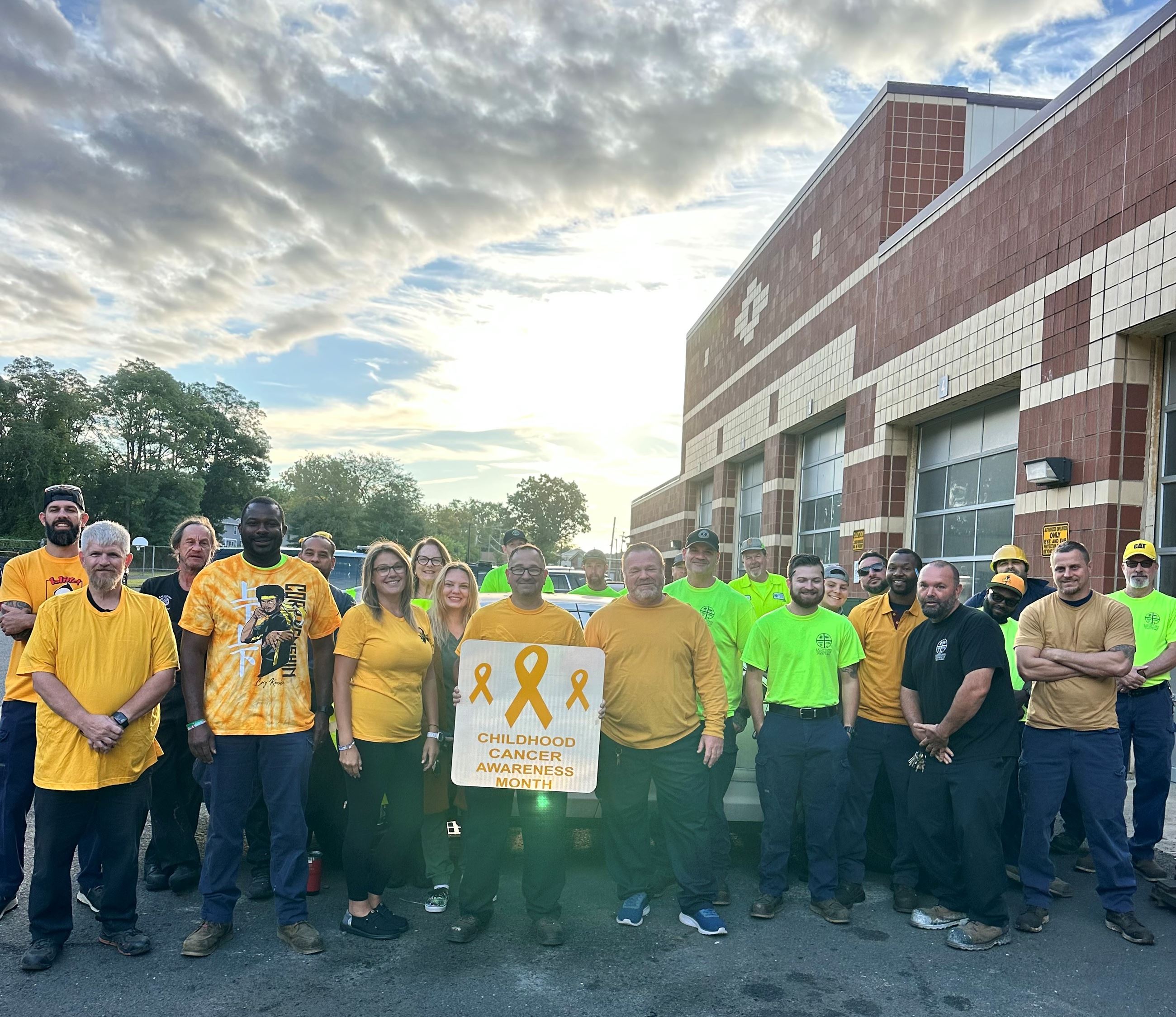 Group of people outside a brick building holding a sign reading “Childhood Cancer Awareness Month” with gold awareness ribbons.